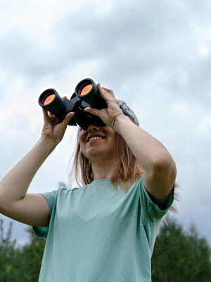 smiling young blonde woman bird watcher in cap looking through binoculars at cloudy sky in forest